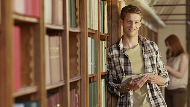 一个年轻人拿着平板电脑站在图书馆书架旁的肖像视频素材