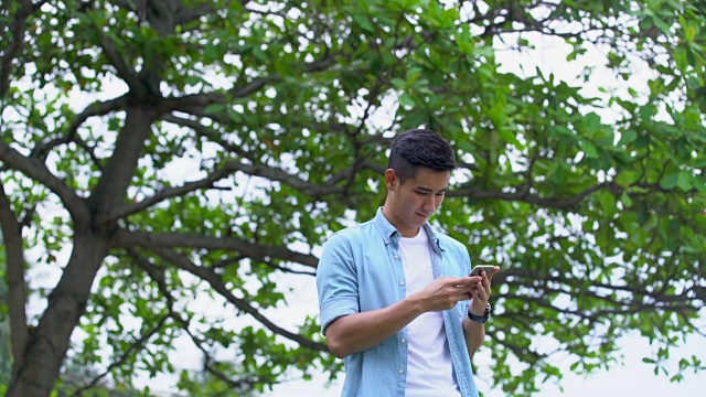 一个年轻人在树下用手机发短信。视频素材