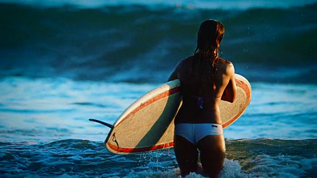 在加州圣地亚哥，一个可爱的女孩背着她的冲浪板在夏季日落的海洋中捕捉更多的海浪。视频下载