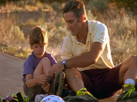 父亲+儿子坐在地上/父亲帮助儿子调整直排轮滑视频素材