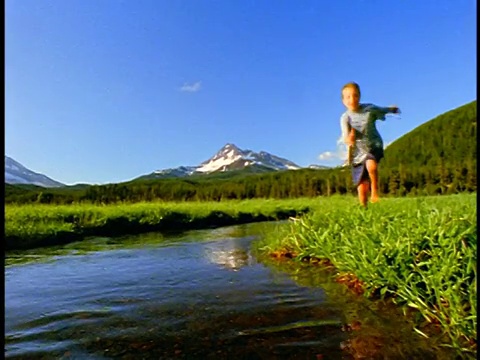 慢镜头男孩跳进溪流向相机+溅/火花草地，喀斯喀特山脉，俄勒冈州视频下载