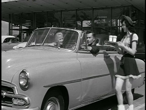1952年，女服务员穿着牛仔装在免下车餐厅的敞篷车里给一对夫妇端盘子视频素材