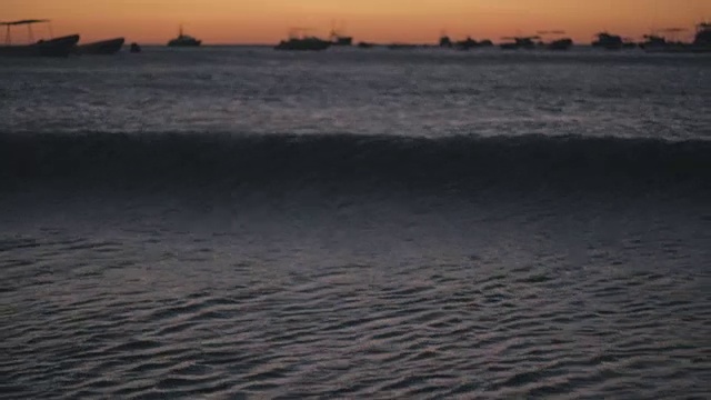 从低角度看太平洋的黄昏。船在背景上散焦。橙色和蓝色是美丽的颜色。向摄像机走来的波浪。视频下载