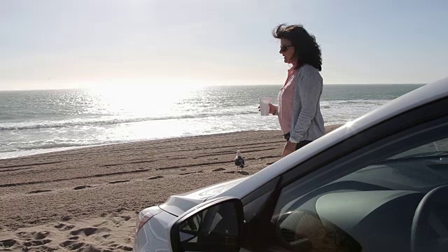 女人端着咖啡走到车前，望着大海。视频素材