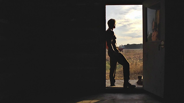 一个体格健壮的老式农场工人站在一间小木屋的门口，看着日落。视频素材