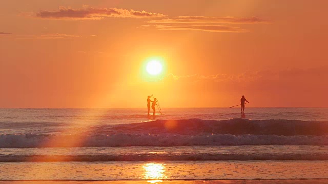 3个站立的冲浪运动员，海浪，橙色的太阳升起的剪影视频下载