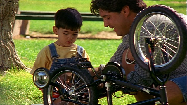 慢镜头摄影机拍摄了一名西班牙男子向小男孩展示如何在户外修理自行车视频素材