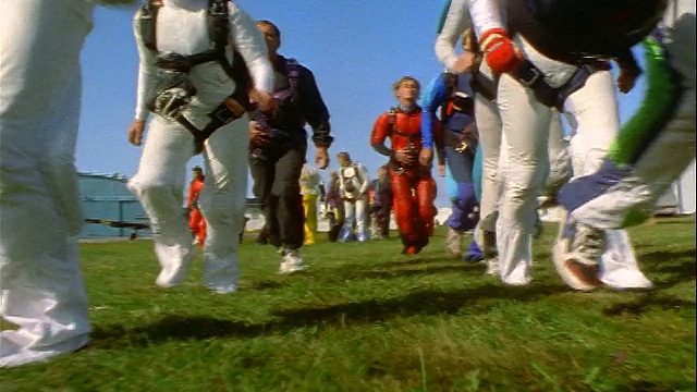 低角度的视角，两行跳伞者穿着五颜六色的工作服在草地上慢跑经过相机/瑞典视频下载