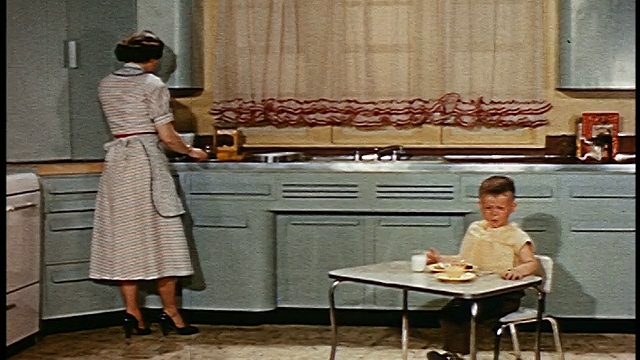 1952年，年轻男孩在厨房的小桌子上吃饭，而妇女在柜台工作视频素材