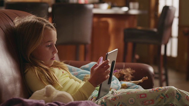 小女孩穿着睡衣舒适地躺在沙发上玩iPad;职业妈妈在背景中交叉。视频素材