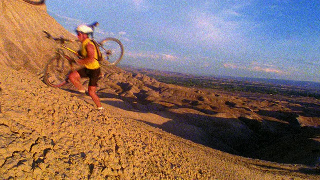在沙漠中，两名男性山地自行车手扛着自行车往山上跑。科罗拉多视频素材