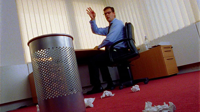 倾斜的低角度商人在办公桌上把皱巴巴的纸扔到地板上，在前景被垃圾桶扔得凌乱不堪视频下载