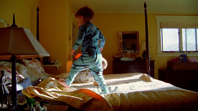慢镜头拍摄周围两个金发的蹒跚学步的男孩穿着睡衣走路，摔倒+在大床上玩耍视频素材