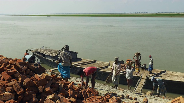 人们从河岸上往一艘传统的木船上装载砖块和压舱物，身后是平坦开阔的河流视频下载