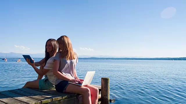WS ZI两个年轻的女人在湖边的甲板上用笔记本电脑和平板电脑工作视频下载