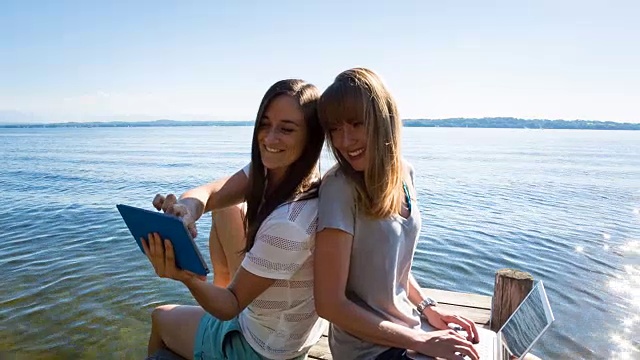 ZI女士是两个年轻的女人，在湖边的甲板上用笔记本电脑和平板电脑工作视频素材