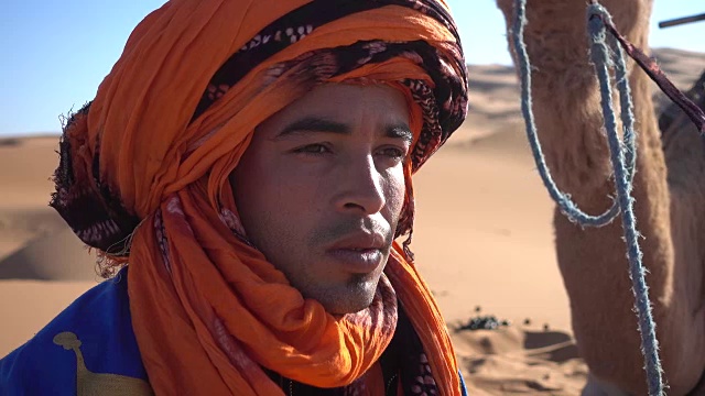 在沙漠中，一位身穿传统头巾的男子在他的骆驼旁边休息视频下载
