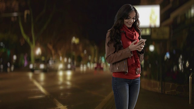 年轻女子晚上在街上用智能手机，一边笑一边发短信视频素材