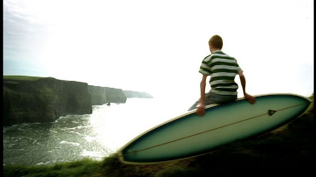 高对比度的后视镜摄影男孩坐在悬崖边缘的冲浪板上，海洋/悬崖的莫赫，爱尔兰视频素材