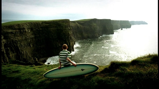 高对比度后视图倾斜男孩坐在悬崖边缘的冲浪板上，爱尔兰莫赫悬崖视频素材