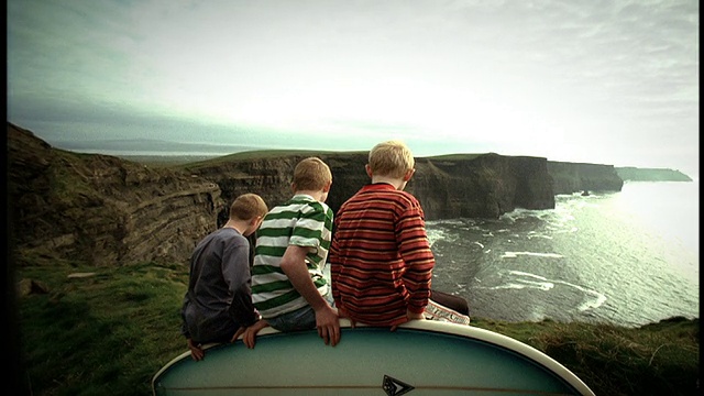 后视图，3个男孩坐在冲浪板上点头悬崖/莫赫悬崖，爱尔兰视频素材
