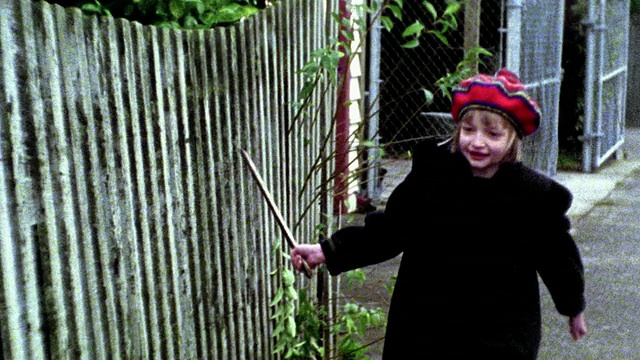MS年轻的金发女孩在红帽子跑向相机旁边的栅栏拖着棍子沿着它视频素材