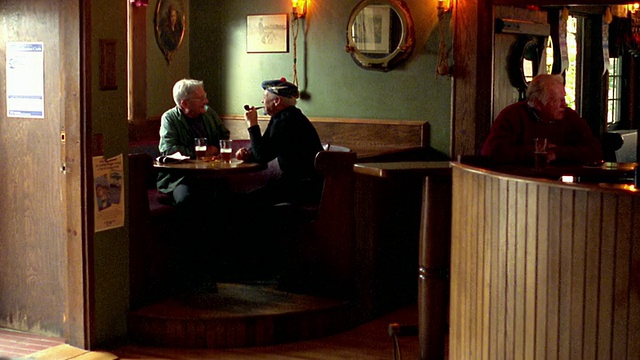 (广角镜头)2名资深男子坐在酒吧桌旁，拿着啤酒杯/ 1名抽烟斗/第三名男子在酒吧/新斯科舍省视频素材
