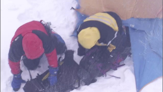 在一场暴风雪中，两名成年人从一个帐篷里爬出来，帐篷在狂风中摇摆着，他们在捡背包。视频下载