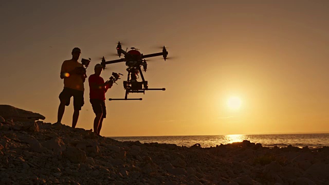 日落时分，两名男子拿着无人机的遥控器在海滩上降落视频下载