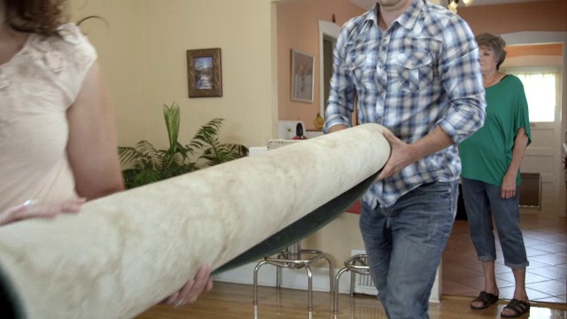 年轻的男人和女人拿着卷起来的地毯在老年妇女的家里。视频下载
