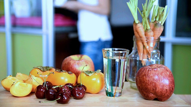 厨房的木桌上放着各种水果、水杯，背景是女人模糊的视频素材