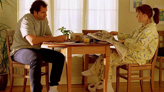 一对夫妇坐在厨房的桌子上，女人在看报纸，男人看起来很沮丧视频素材