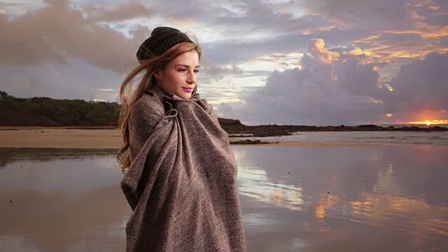 一名20多岁的年轻女子站在户外的海滩边，手里拿着一条毯子取暖视频下载