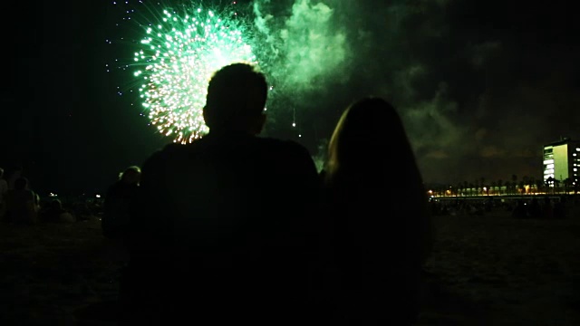 一对相爱的夫妇的剪影坐在巴塞罗那的海滩上凝视着夜晚的烟花与城市天际线为commerce节日。视频素材