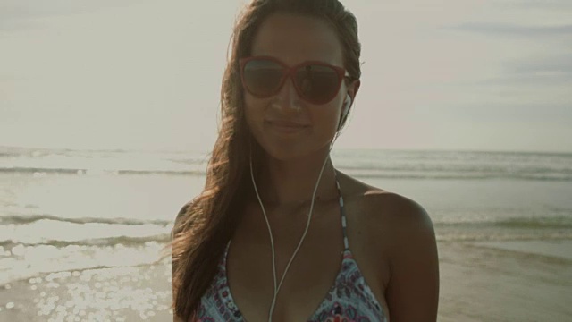 在法国南部的大西洋海滩上，一名身穿比基尼、戴着耳机和太阳镜的美女在镜头前微笑视频下载
