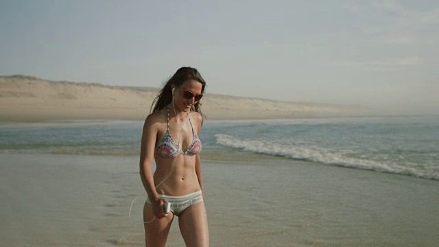 在法国南部荒凉的大西洋海滩上，穿着比基尼戴着耳机的美丽女子视频下载