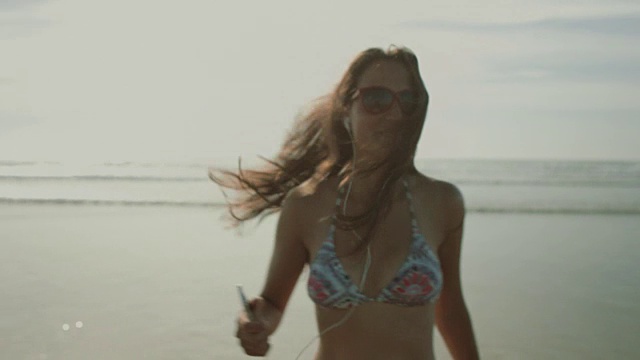 在法国南部荒凉的大西洋海滩上，穿着耳机和比基尼，走在一个转身微笑的美女身后视频下载
