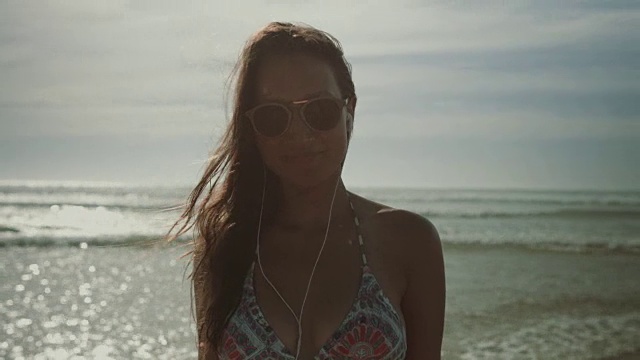 在法国南部的大西洋海滩上，一位戴着太阳镜和耳机的比基尼美女对着镜头微笑视频下载