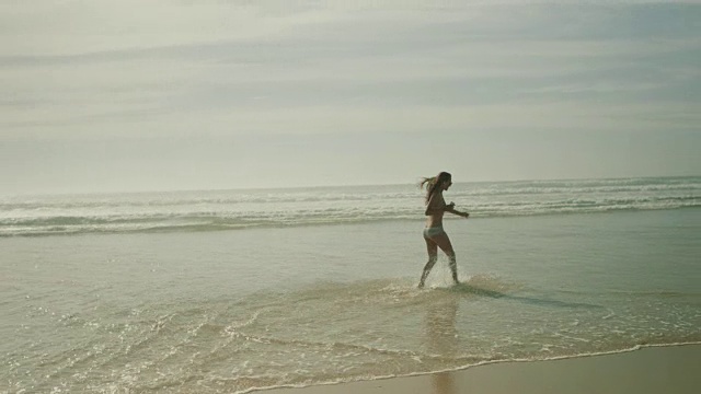 在法国南部荒凉的大西洋海滩上，美丽的女人听着音乐，走着，穿着比基尼在水中跳舞视频下载