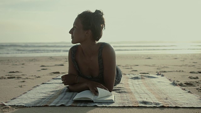 美丽的女人环顾四周，在法国南部大西洋的海滩上，在毯子上看书视频下载