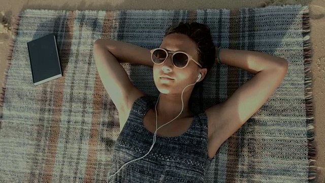 在法国南部的大西洋海滩上，一位戴着耳机坐在毯子上听音乐的美丽女子视频下载