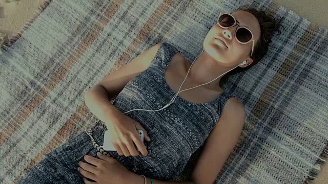 在法国南部的大西洋海滩上，一位戴着耳机坐在毯子上听音乐的美丽女子视频下载