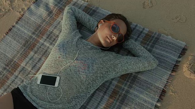 在法国南部的大西洋海滩上，一位美丽的女子坐在毯子上，戴着耳机和手机听音乐视频下载