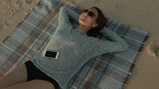 在法国南部的大西洋海滩上，一名美丽的女子正躺在毯子上，戴着耳机和移动设备听音乐视频下载