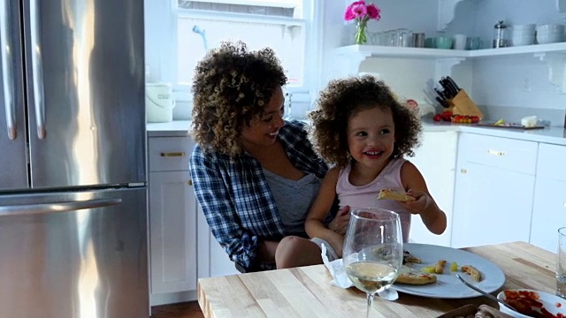 在厨房里，微笑的小女孩坐在妈妈的腿上吃披萨视频下载