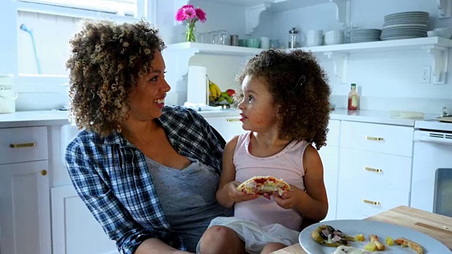 微笑的小女孩坐在厨房妈妈的膝上吃披萨视频下载