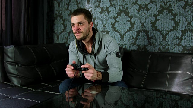 一个年轻的成年人坐在沙发上，用游戏手柄或操纵杆玩视频游戏的慢动作镜头视频下载
