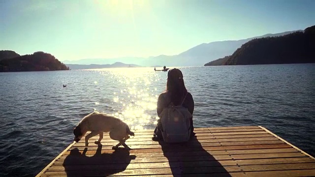 年轻女子与狗坐在湖边的码头上休息视频素材