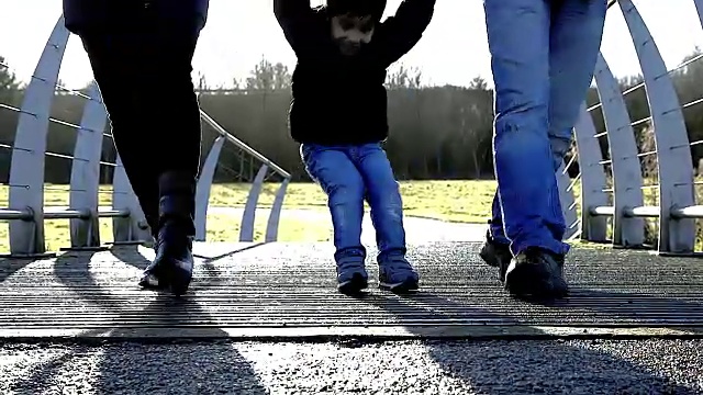 妈妈和爸爸走在公园的桥上，小儿子在他们之间摇摆。视频下载
