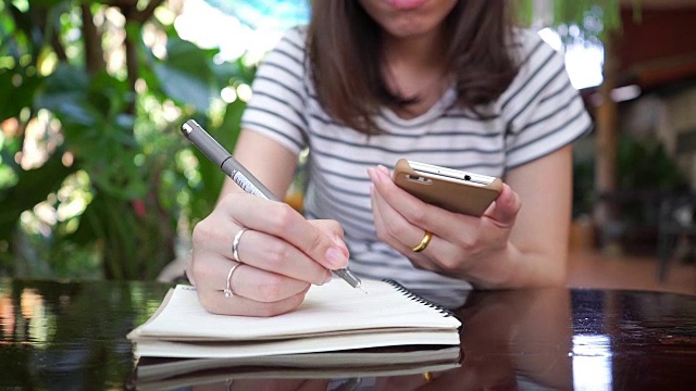 年轻女子一边用智能手机一边在笔记本上写字。视频素材